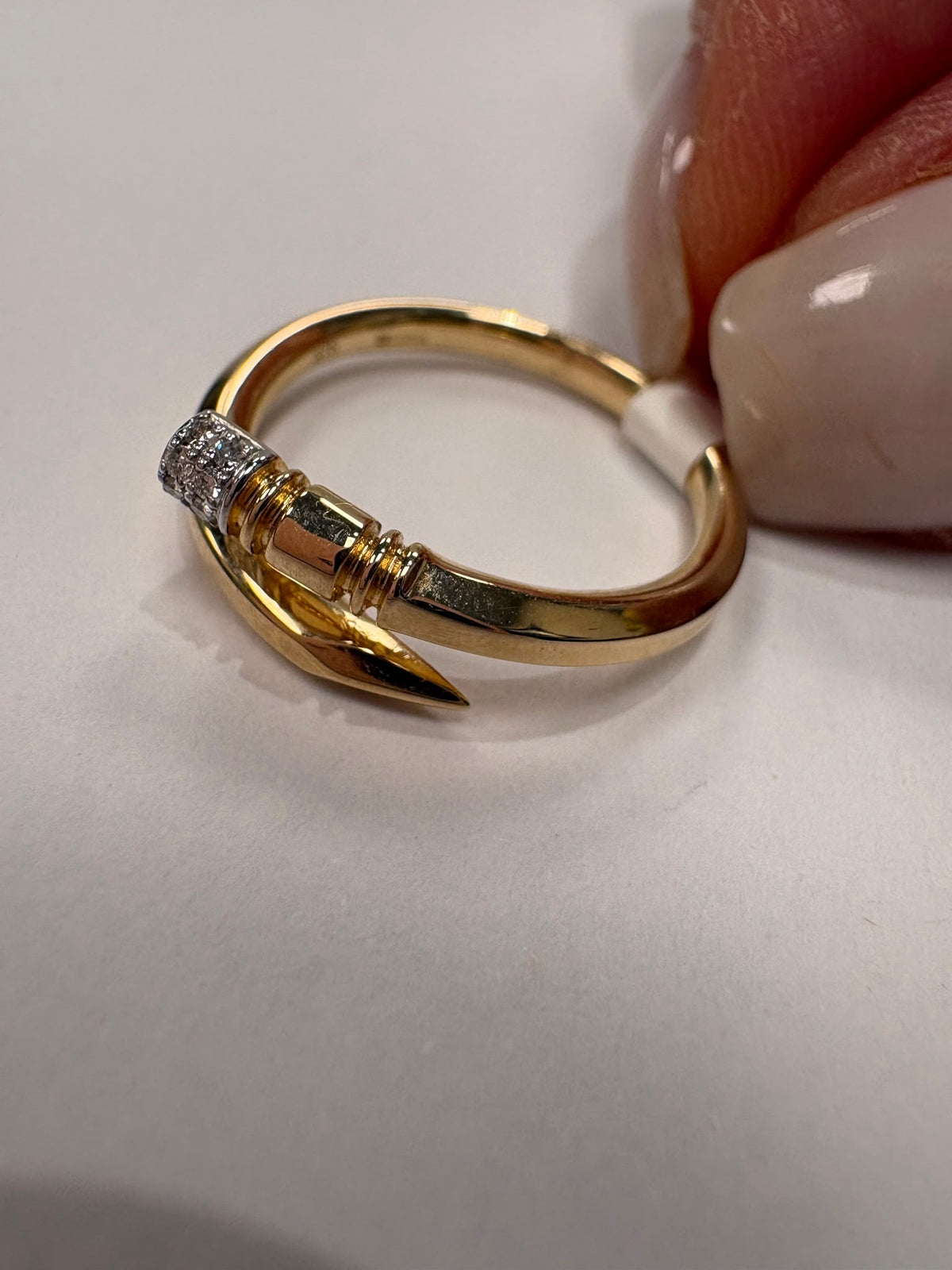 A gold ring designed like a pencil with a small clear gemstone cluster on the side, held by fingers against a plain light background.