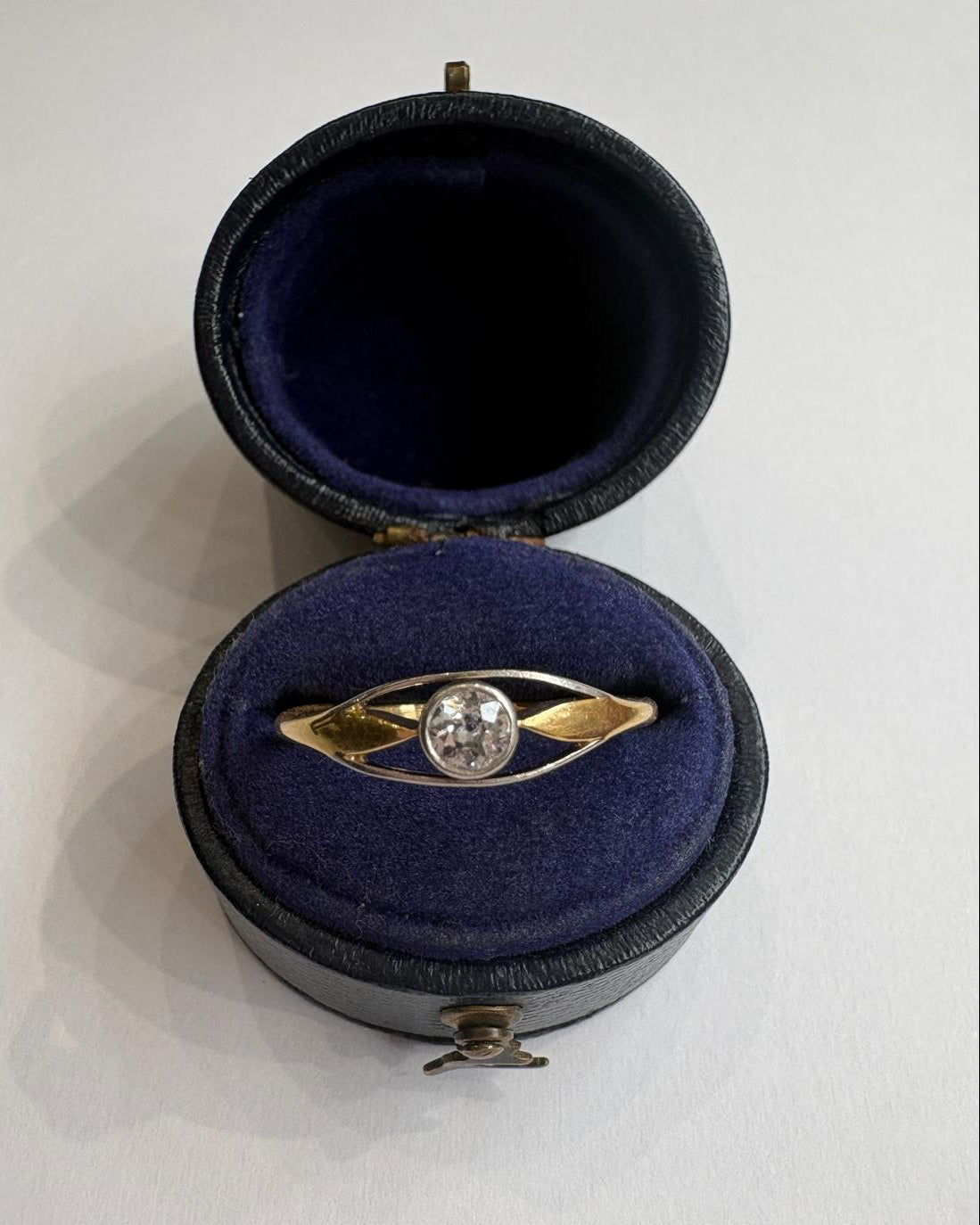 A vintage gold ring with a clear round gemstone, displayed inside an open round black velvet jewelry box on a white background.