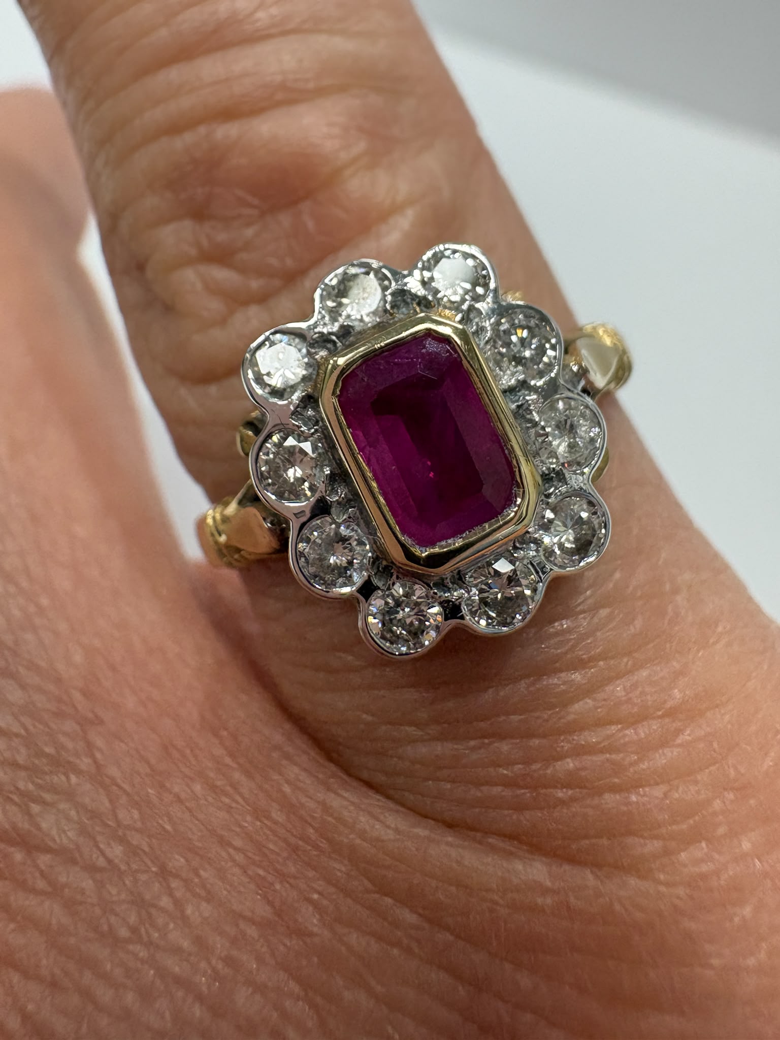 A vintage-style ring with a rectangular emerald-cut pinkish-red center stone surrounded by small clear stones in a silver-toned setting, worn on a finger. The band appears gold-toned on one side and silver-toned on the other.