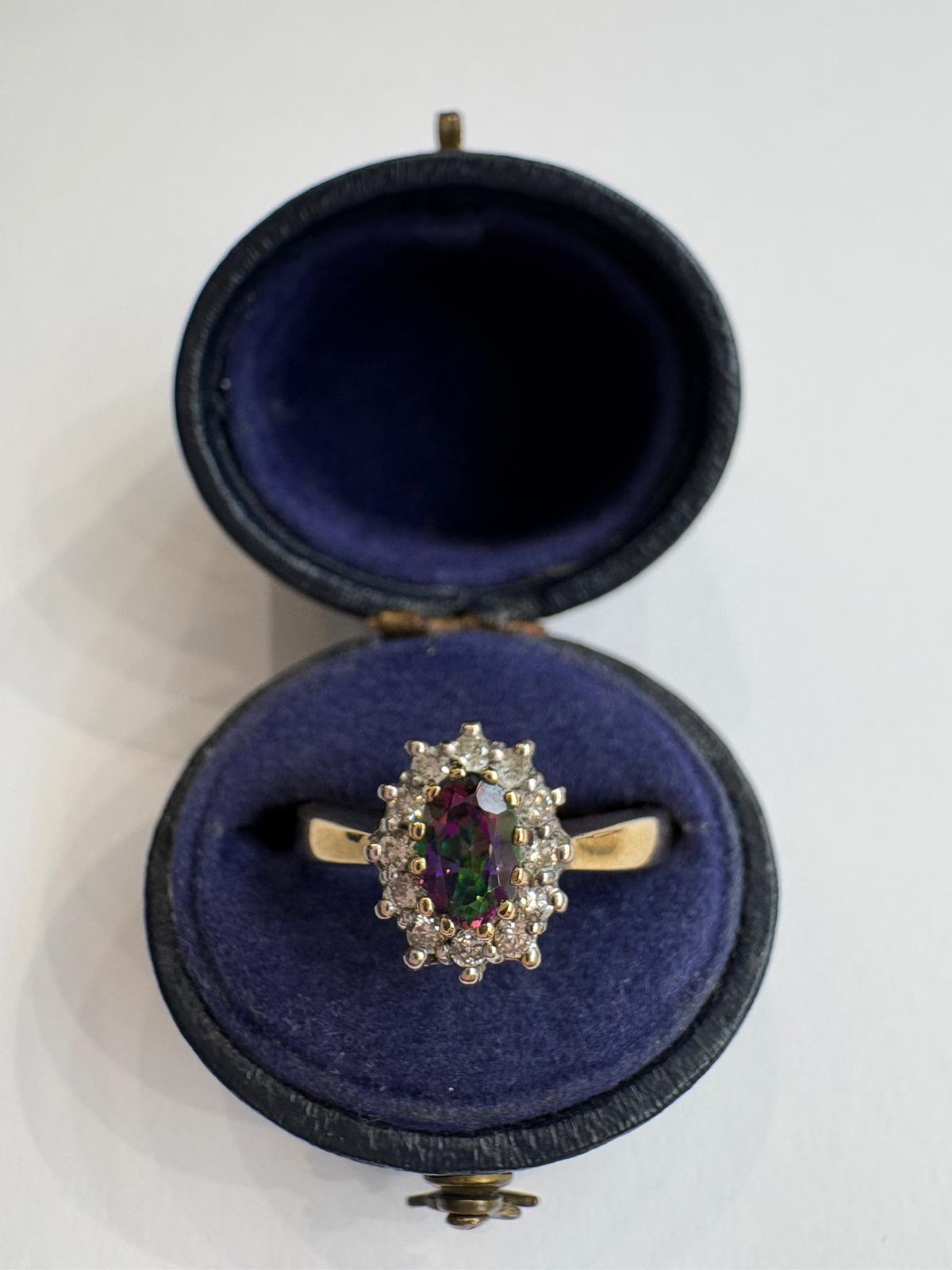 A yellow gold ring with a central oval mystic topaz, surrounded by diamonds, displayed inside an open dark blue velvet ring box on a white background.