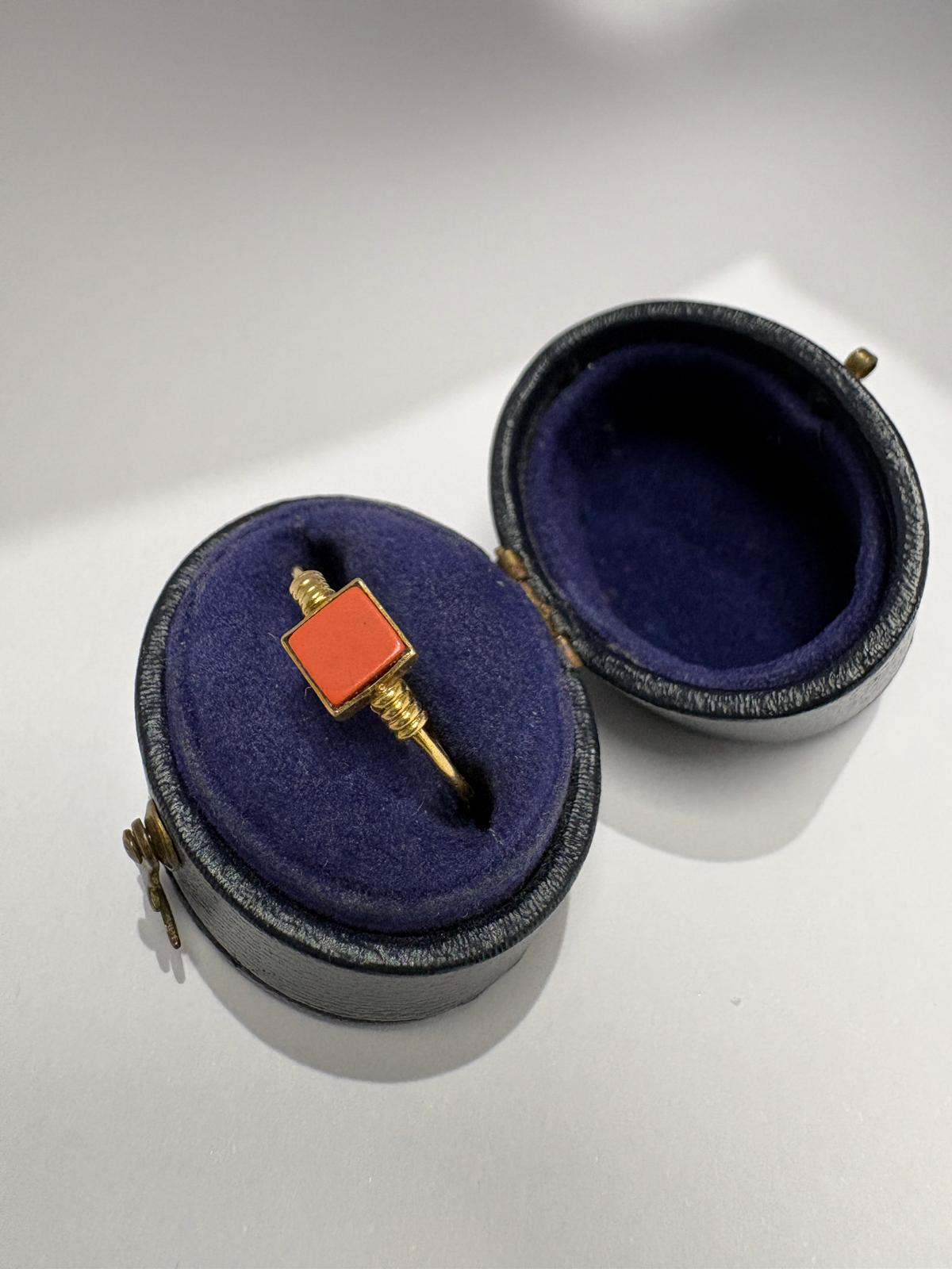 Angled view of a pre-loved 14ct yellow gold natural coral dress ring resting in an open antique navy blue leather ring box. The vivid salmon-orange square coral tablet and gold granulation shoulder detail are clearly visible against the blue velvet cushion.