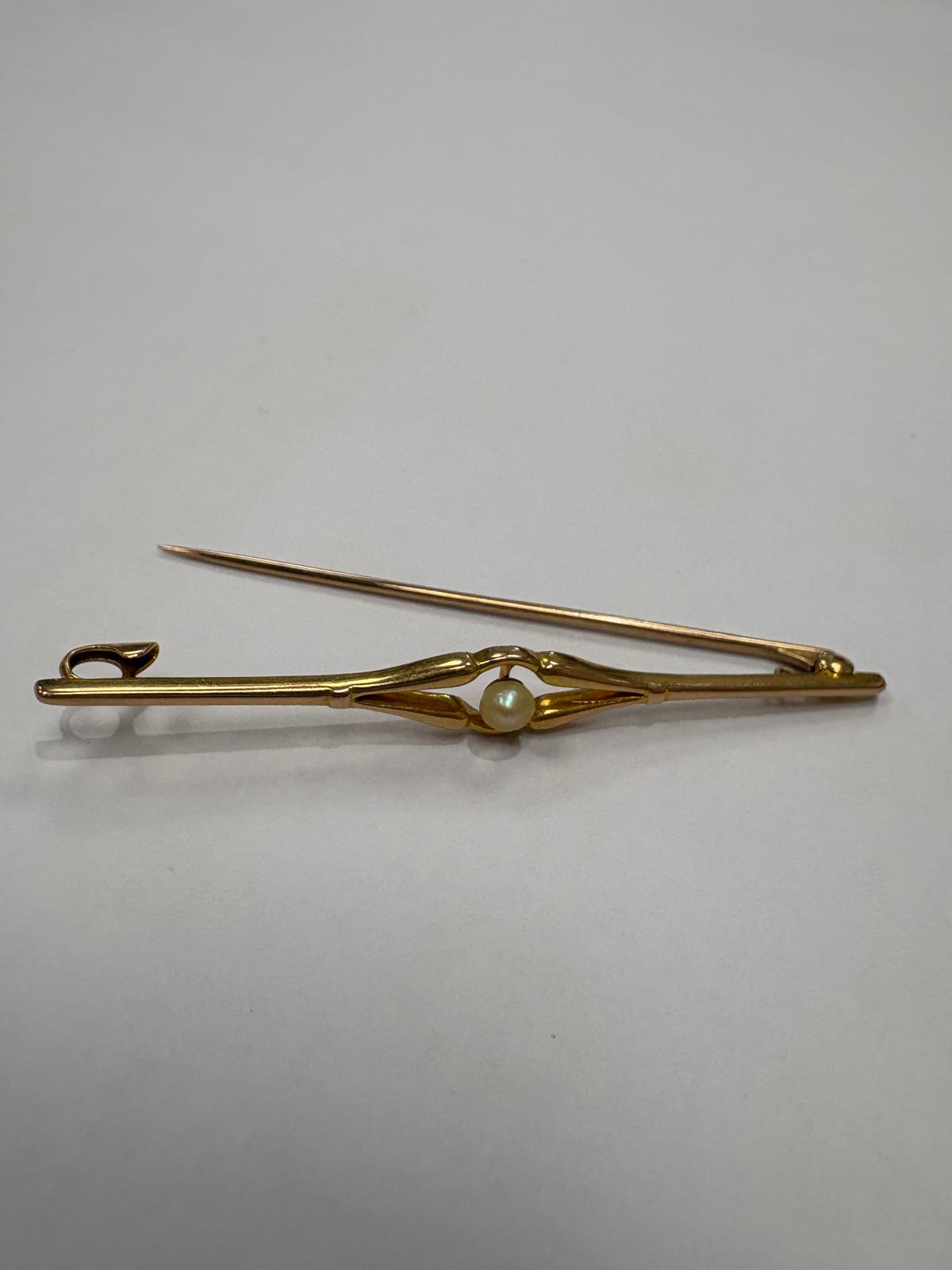Slightly angled front view of a pre-loved Edwardian 18ct yellow gold natural pearl bar brooch on a white surface. The elegant open scroll centrepiece cradles a single natural pearl, the long tapering gold bar extending to fine pointed ends on either side.