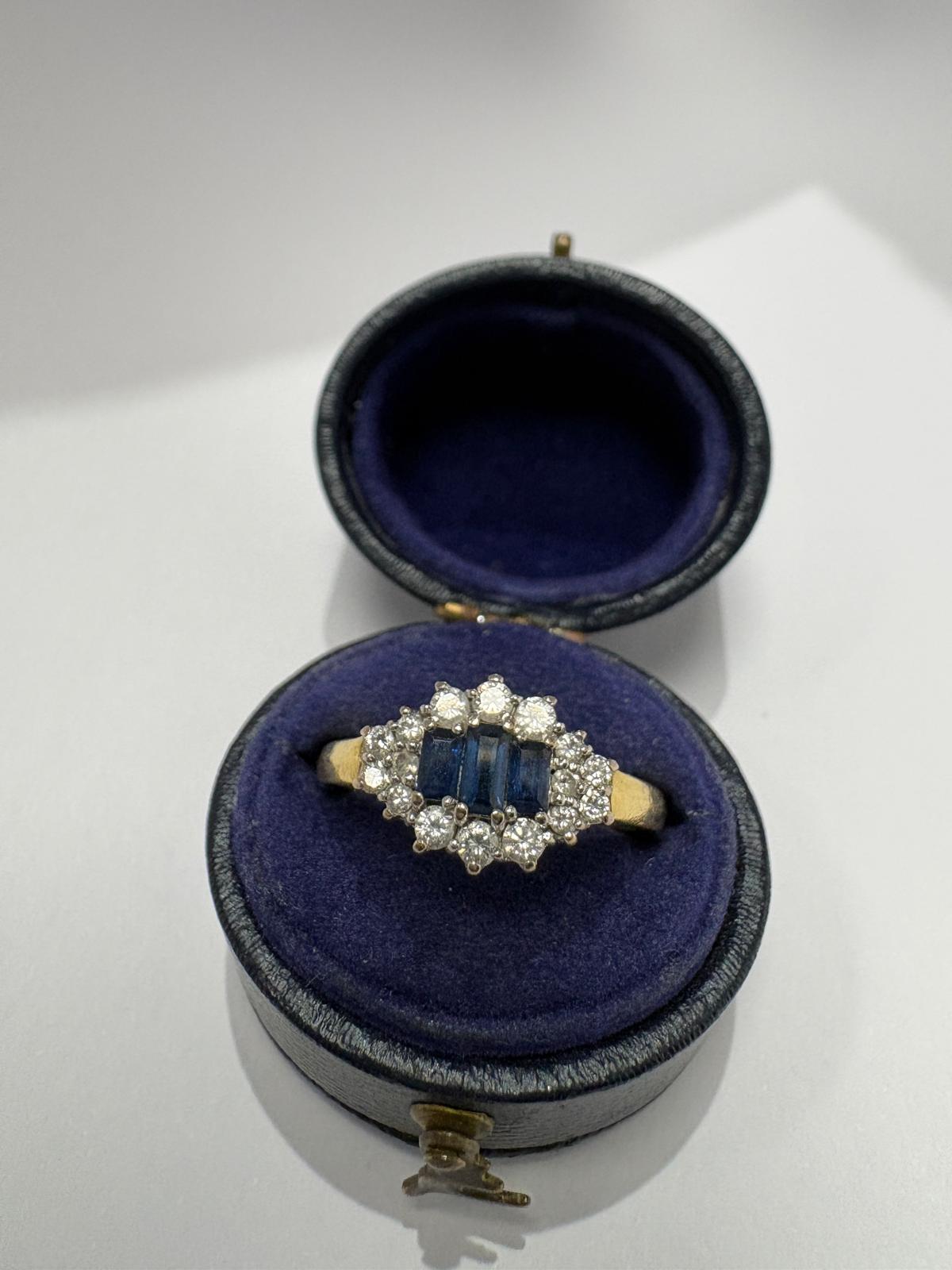 Top view of a pre-loved yellow gold natural sapphire and diamond marquise cluster ring resting on the cushion of an open antique navy blue velvet ring box. Three baguette-cut blue sapphires sit at the centre surrounded by a halo of round brilliant cut diamonds in a marquise-shaped white gold cluster setting.