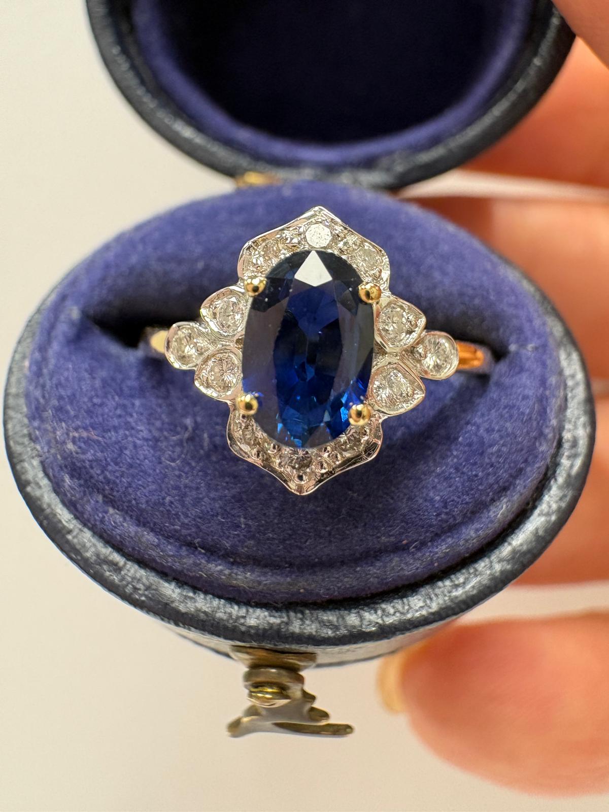 A vintage-style ring with an oval cut blue sapphire and small diamonds in a gold-toned setting, displayed inside an open blue velvet ring box