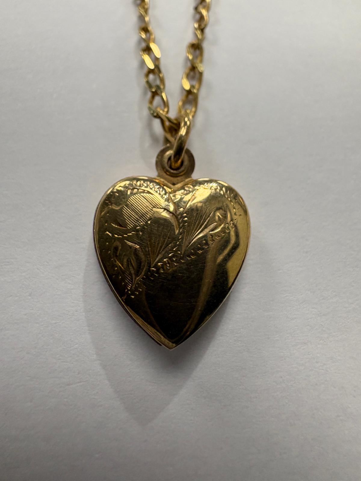Close-up view of the front of a vintage 9ct yellow gold domed heart locket from a slightly lower angle. The hand-engraved foliate decoration is clearly visible across the surface, with fine line and leaf motifs. The jump ring bail and fine oval link chain are visible at the top.