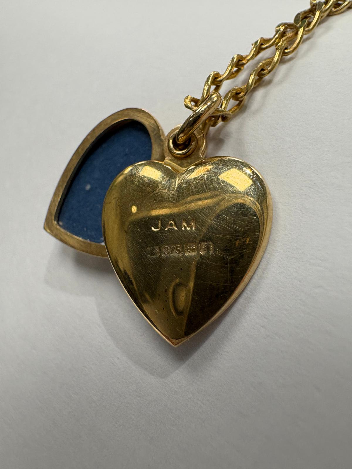 Close-up of the reverse of a vintage 9ct yellow gold heart locket showing the plain polished back. British hallmarks are clearly visible including the 375 gold purity mark, along with maker's initials JAM. The locket is shown open with the blue fabric interior of the back panel visible to the left.