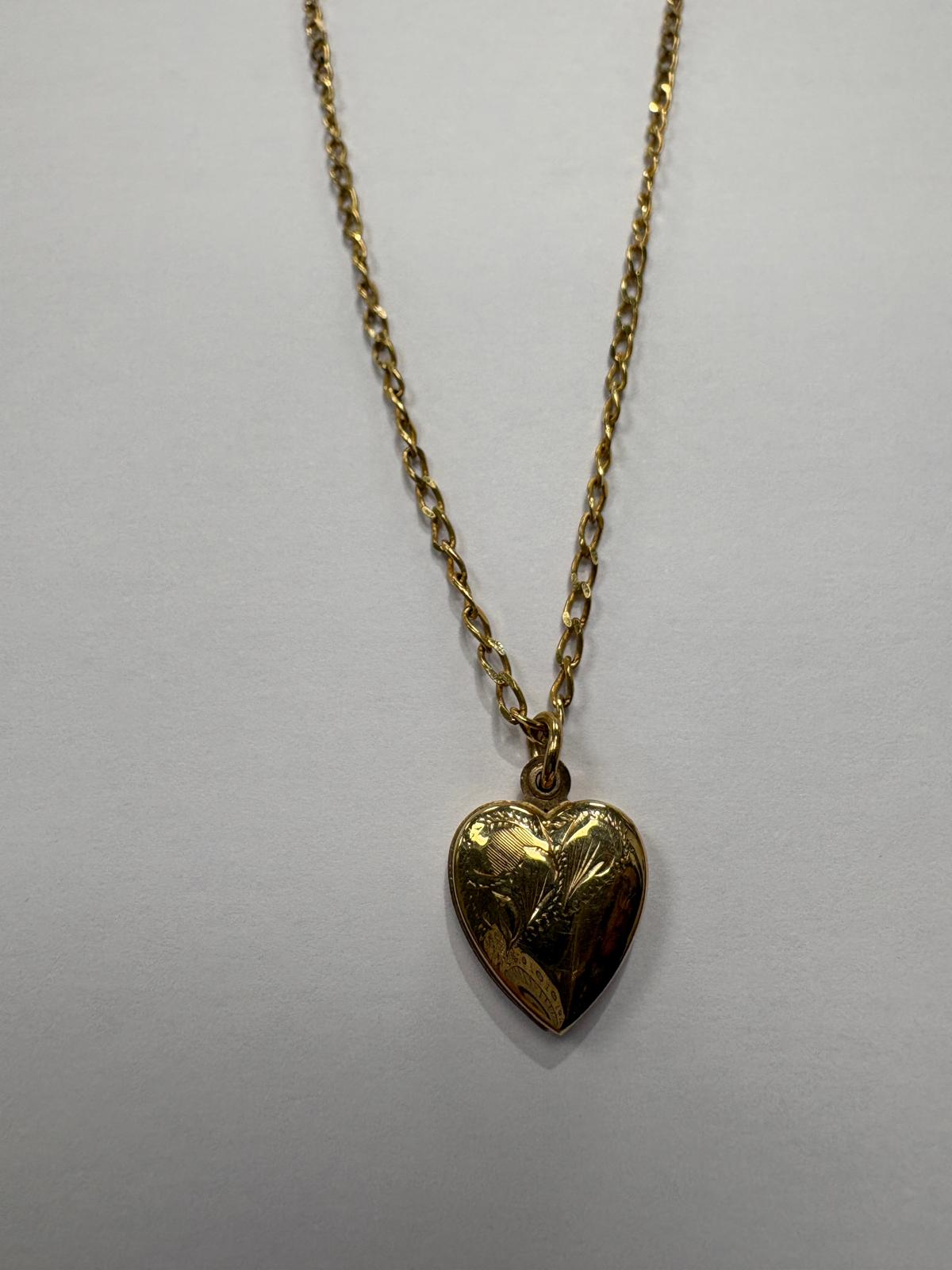 Full length view of a vintage 9ct yellow gold heart locket necklace laid flat on a grey background. A fine yellow gold oval link chain suspends a domed heart-shaped locket with engraved floral and foliate decoration on the front face.