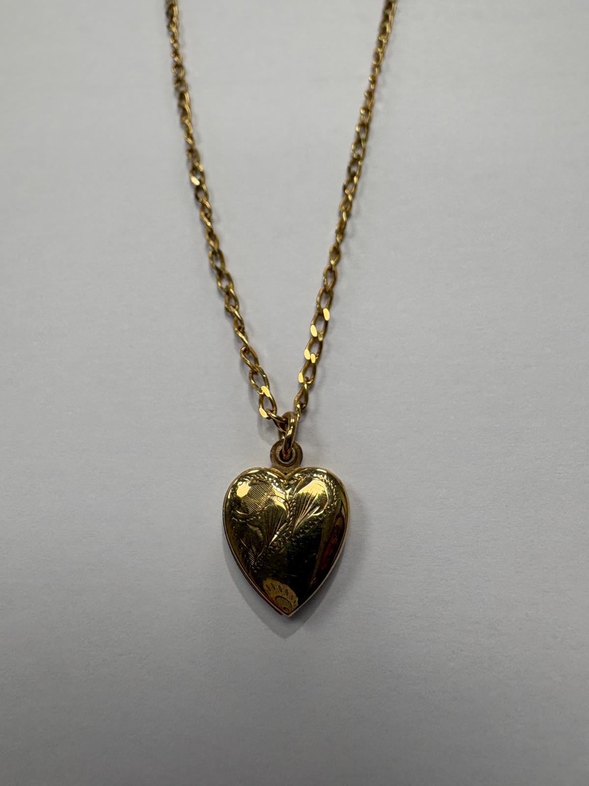 Three-quarter length view of a vintage 9ct yellow gold heart locket necklace on a grey background. The domed heart locket features hand-engraved floral and leaf decoration across the front. A price sticker is visible at the base of the locket. The chain is a fine yellow gold oval link style.
