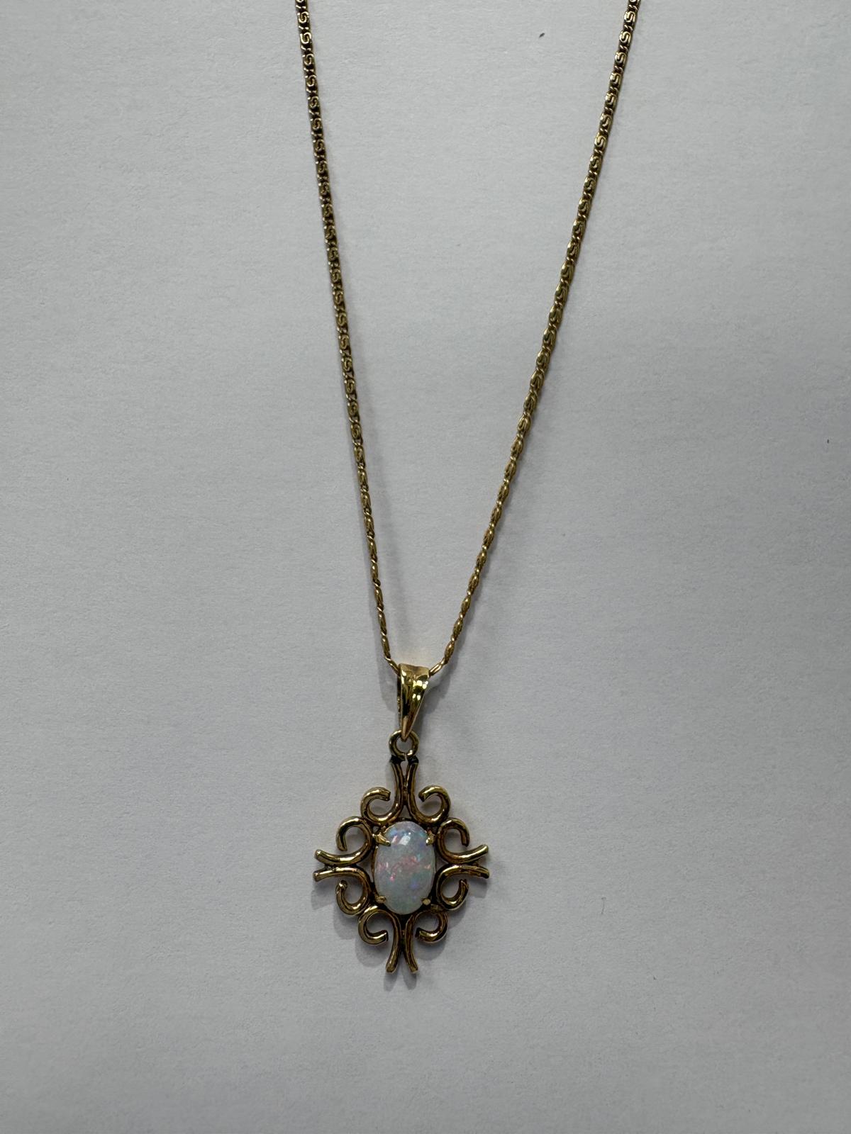 Full length view of a vintage yellow gold necklace laid flat on a grey background. A fine 10k yellow gold fancy link chain suspends a 14ct yellow gold pendant featuring an oval opal cabochon with soft white body colour and pink and blue play-of-colour, held in an ornate open scrollwork setting.