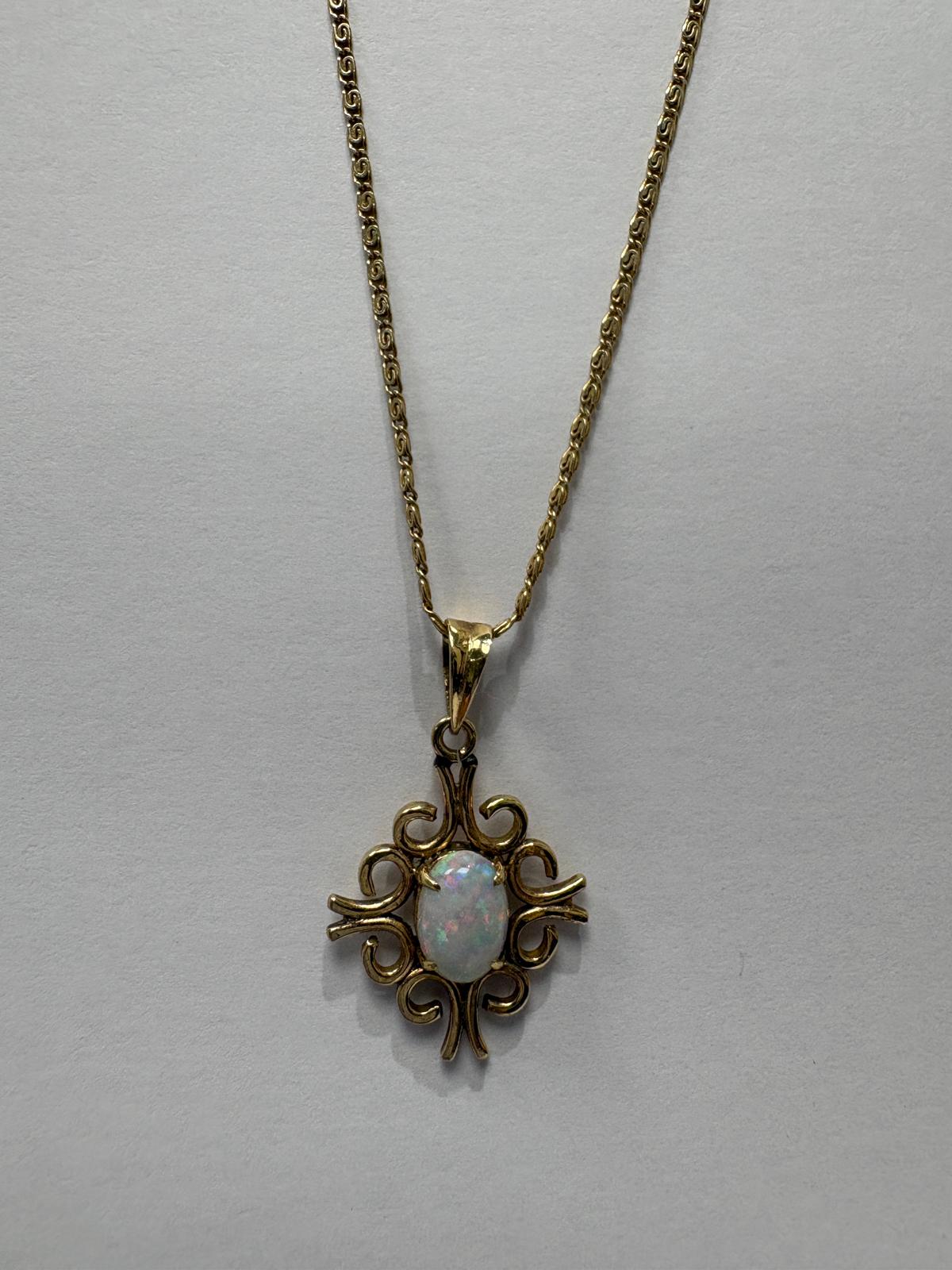 Three-quarter length view of a vintage yellow gold necklace on a grey background. The 10k yellow gold fancy link chain leads to a 14ct yellow gold open scrollwork pendant set with an oval opal cabochon displaying white body colour with flashes of pink, red, and blue play-of-colour.