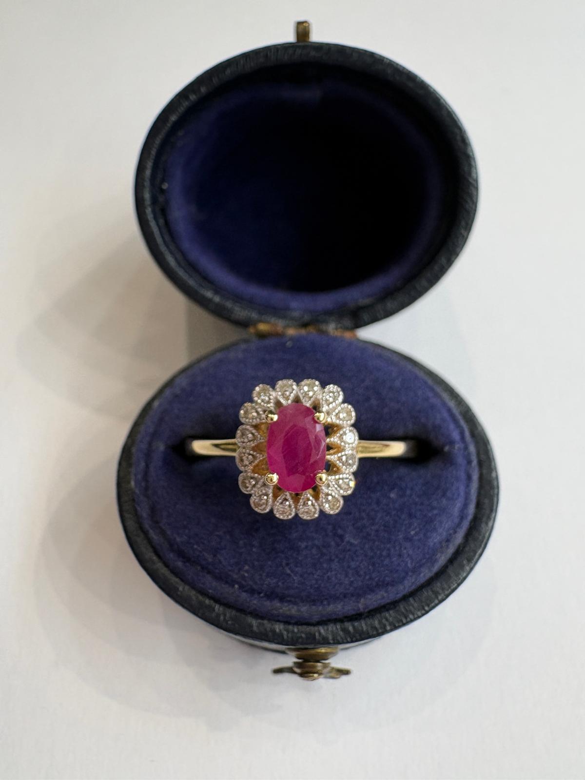 A yellow gold ring with a central oval ruby surrounded by diamonds set as teardrops with milgrain, displayed inside an open dark blue velvet ring box on a white background.