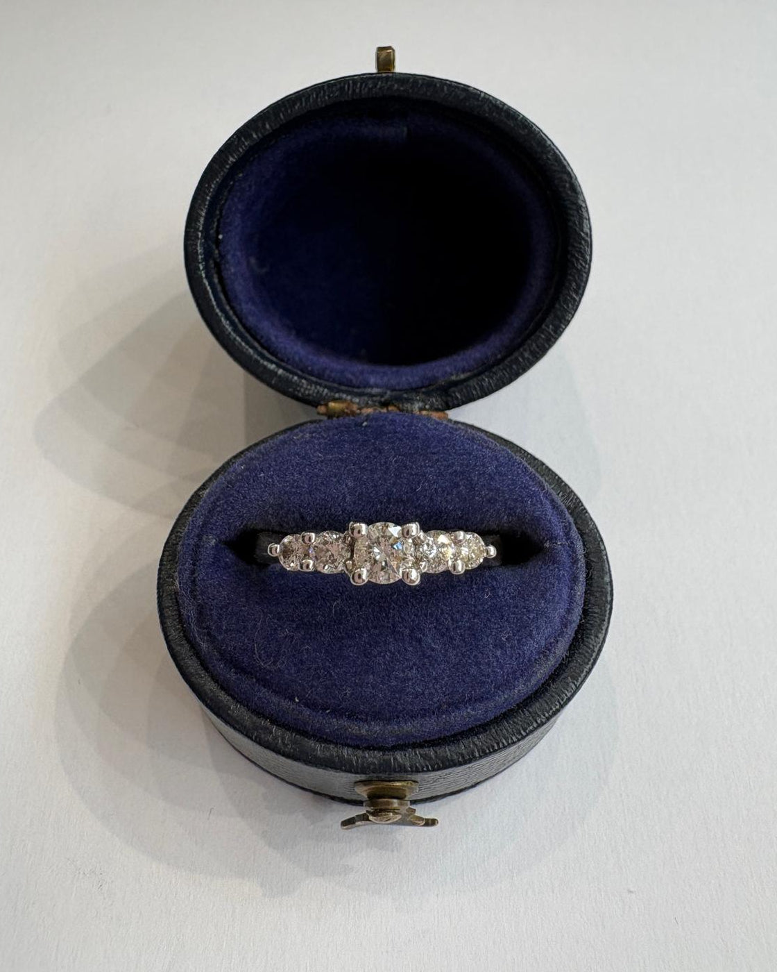 A diamond ring with 5 diamonds, displayed inside an open round presentation box with a dark blue velvet interior on a light gray background.