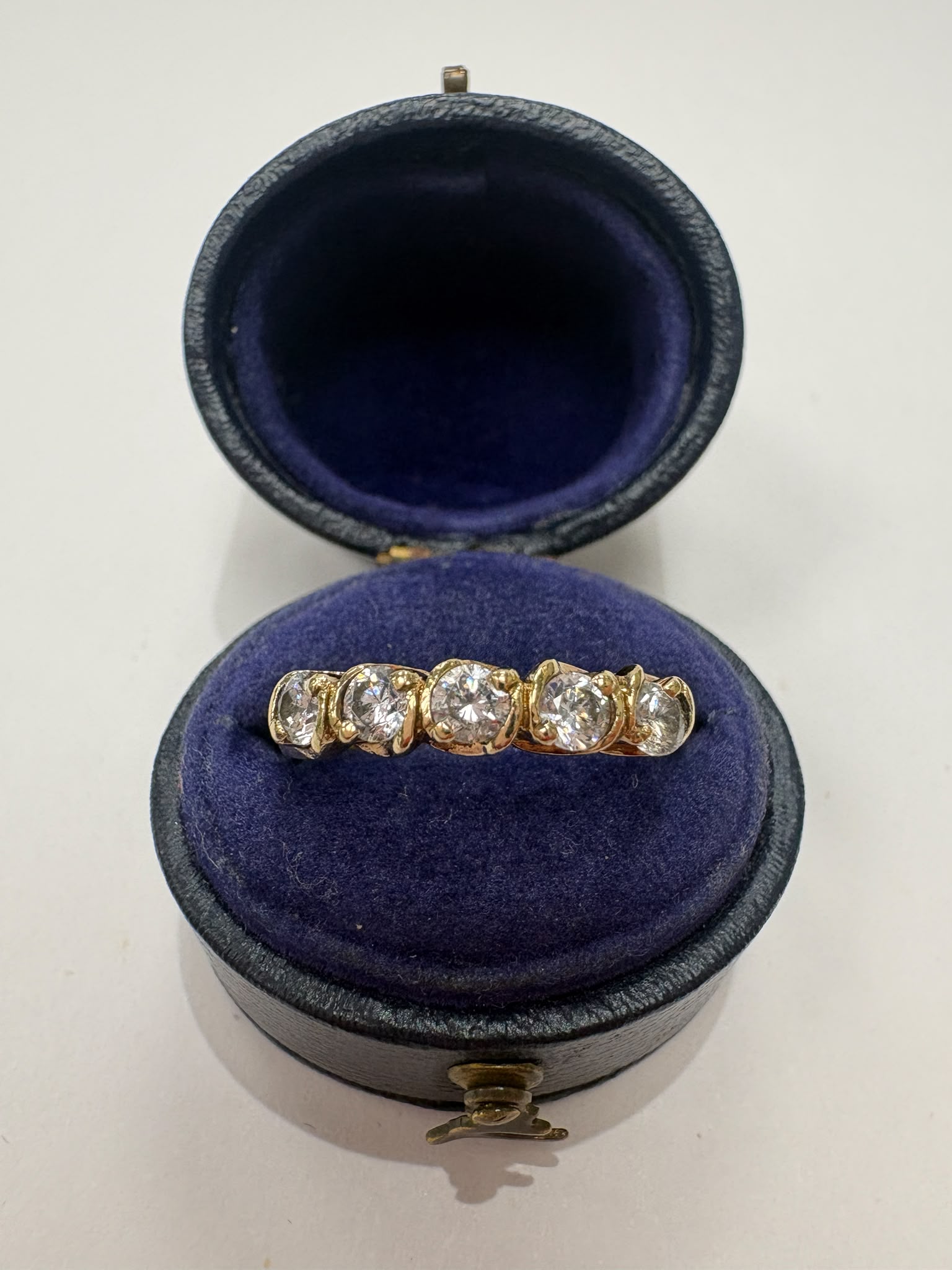A yellow gold five-stone diamond ring displayed inside an open round dark blue velvet ring box on a light gray background.