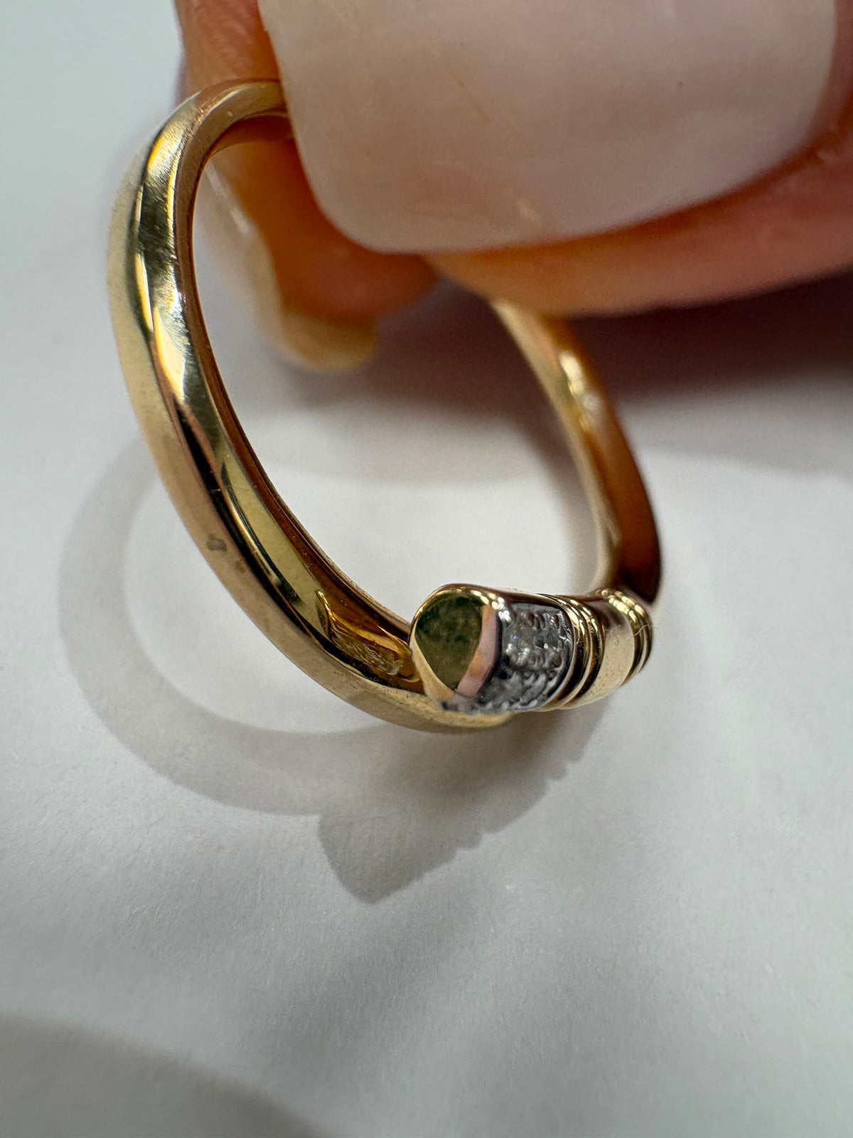 A close-up of a gold ring being held by fingers, featuring a small heart-shaped green gemstone and a row of clear stones on the band.