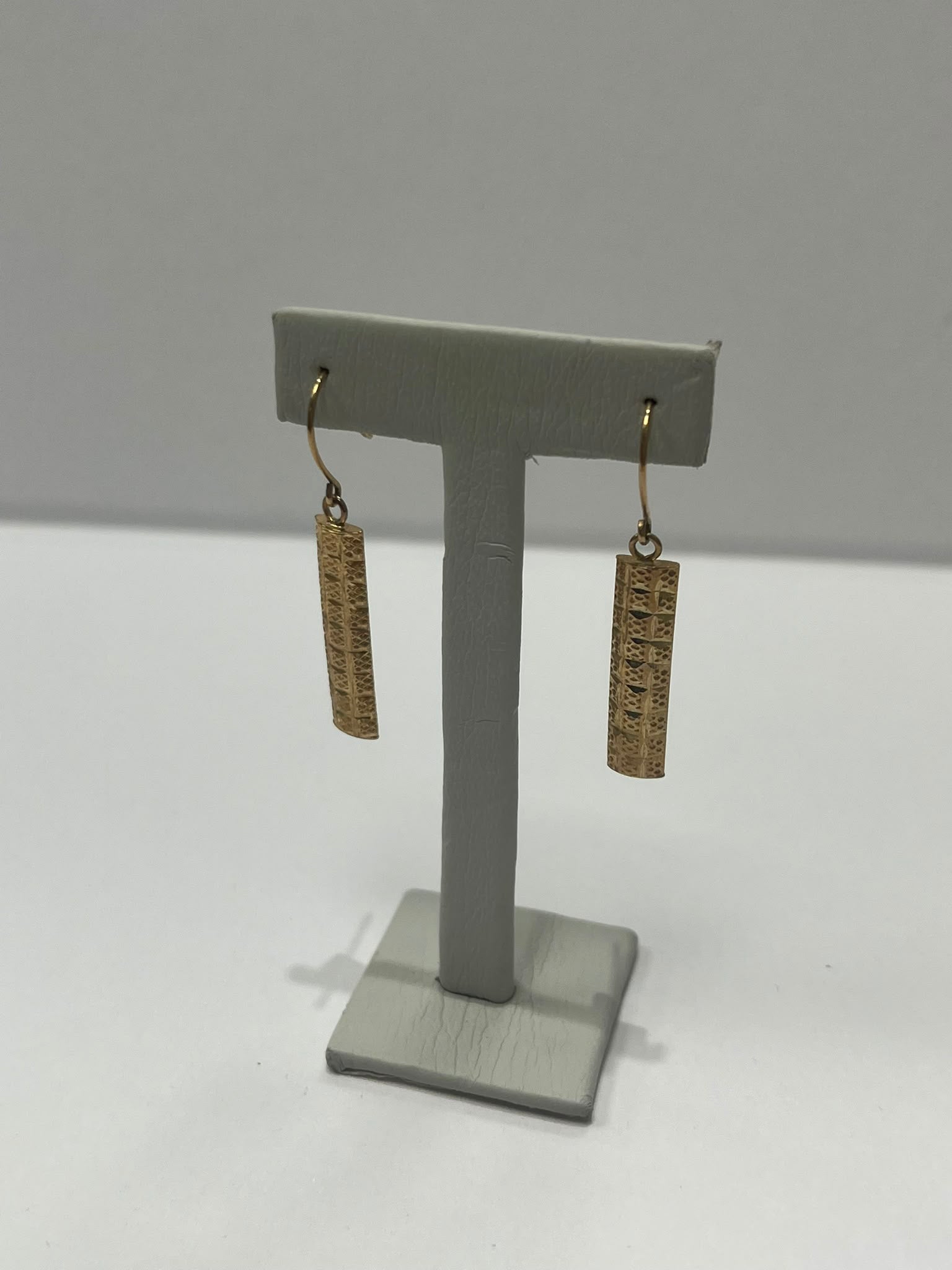 A pair of gold-toned drop earrings with textured rectangular charms, displayed on a gray T-shaped jewelry stand against a plain light gray background.