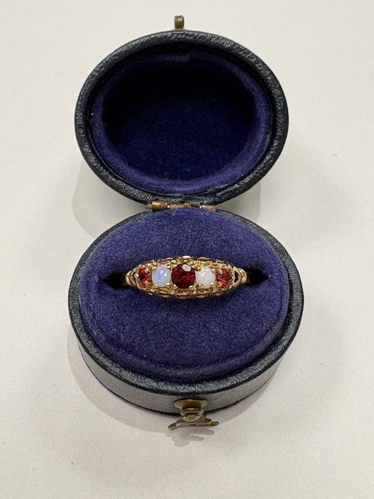 A vintage gold ring with a central red gemstone and white gemstones on either side, displayed inside an open round velvet ring box with a dark purple lining.