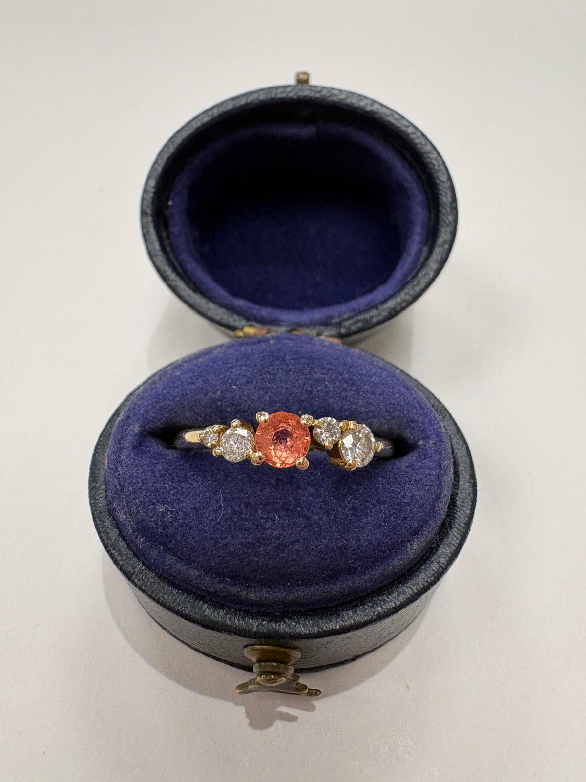 A yellow gold ring with a central round orange sapphire and two small clear stones on either side, displayed inside an open round ring box with a dark blue velvet lining on a light gray background.