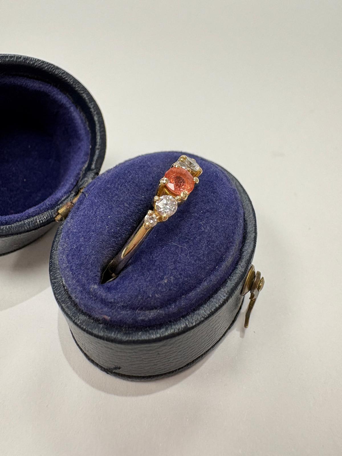 A yellow gold ring with a central orange sapphire and smaller clear accent stones, displayed in an open dark blue velvet ring box
