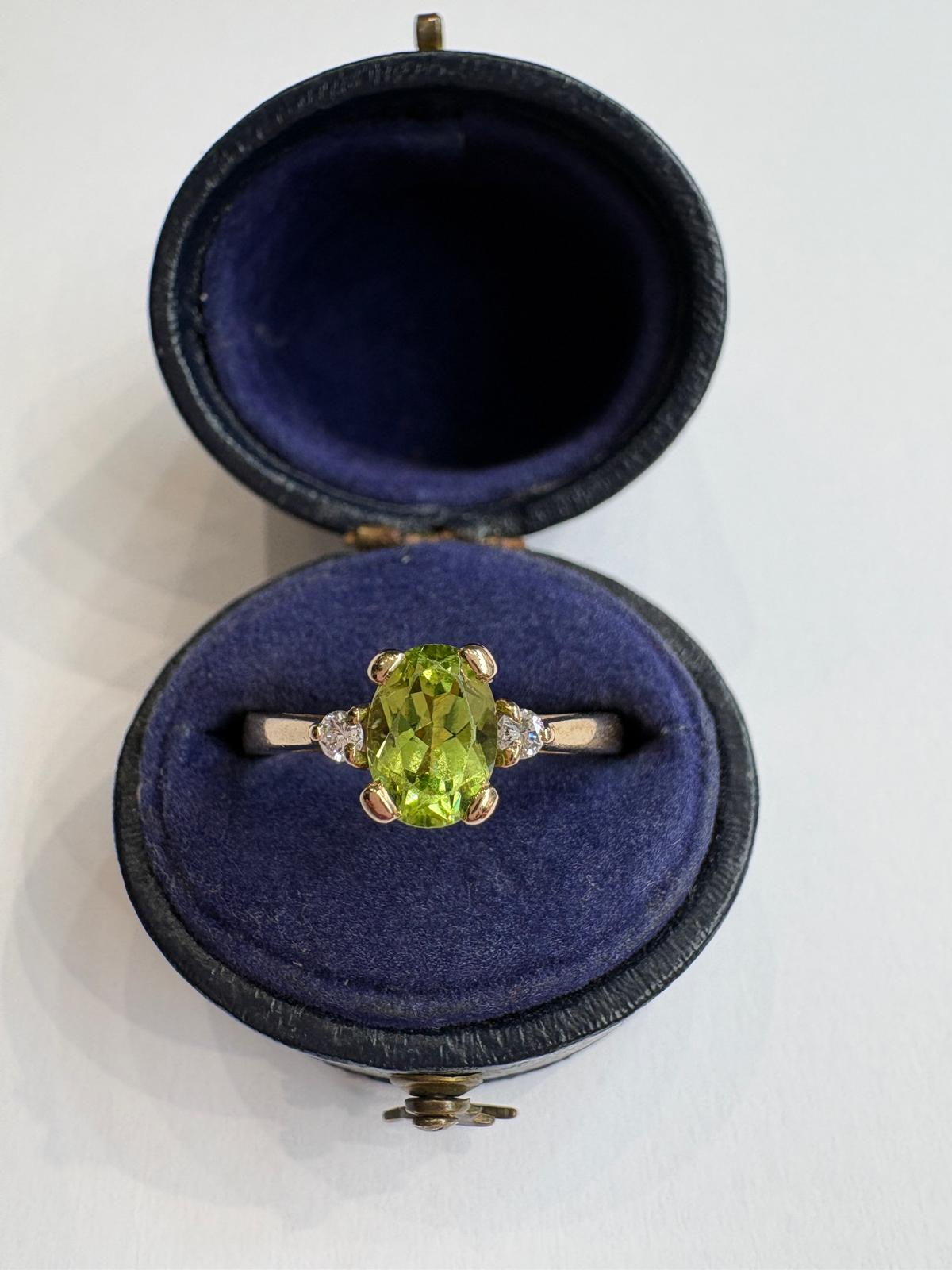 A yellow gold ring with an oval green peridot center stone and small side stones, displayed inside an open dark blue velvet ring box on a white background.