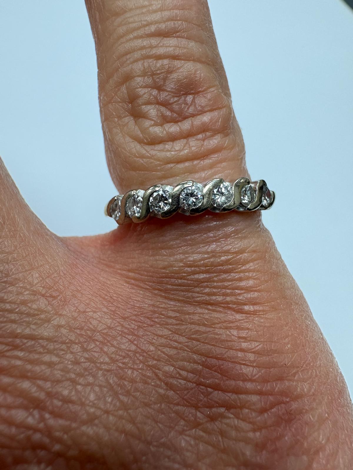 Close-up of a pre-loved yellow gold diamond twist wave eternity ring worn on a finger. The ring features seven round brilliant cut diamonds set in a continuous S-shaped wave white gold setting, each diamond bright and lively. The yellow gold shank is visible beneath the diamond-set face.