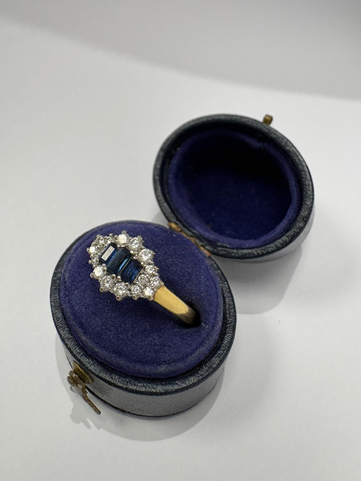 Close-up front face view of a pre-loved yellow gold natural sapphire and diamond marquise cluster ring worn on a finger. Three vivid deep blue baguette-cut natural sapphires sit side by side at the centre, surrounded by a border of round brilliant cut diamonds in a claw-set white gold marquise cluster setting. The yellow gold shank is visible at the sides.