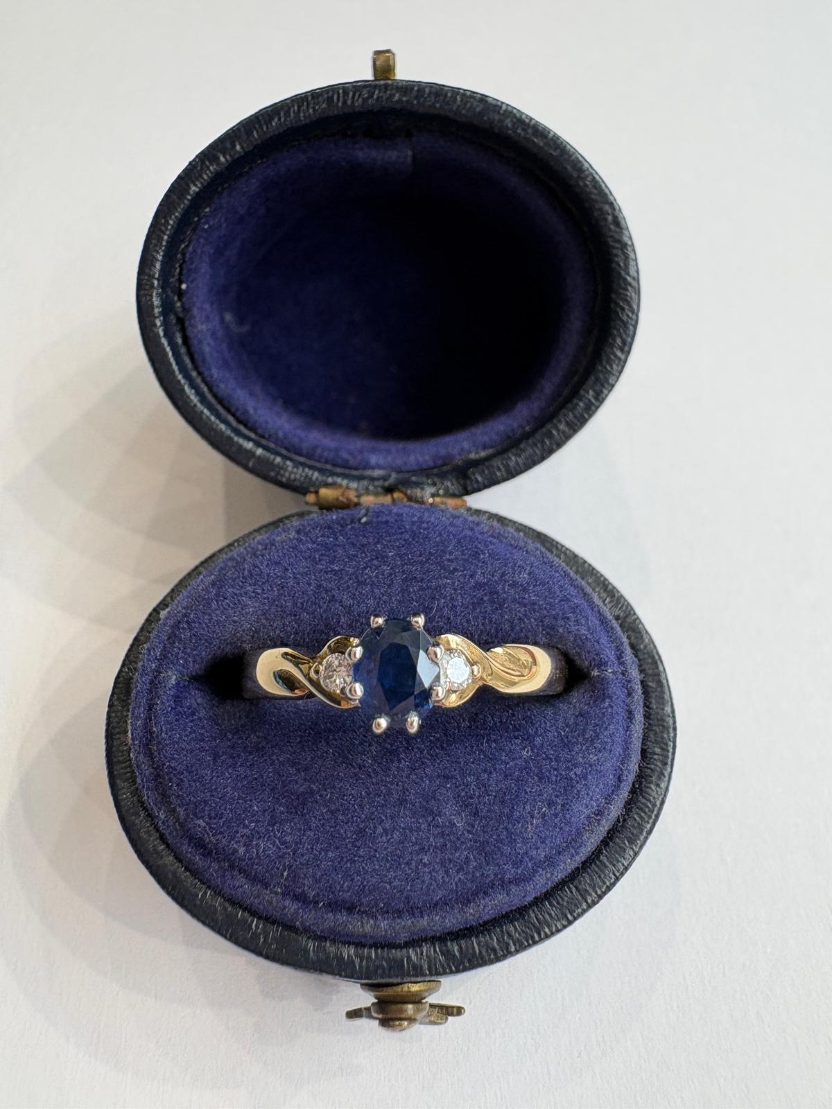 A gold ring with a central oval periwinkle sapphire and small diamonds, displayed in an open round dark blue velvet ring box on a white background