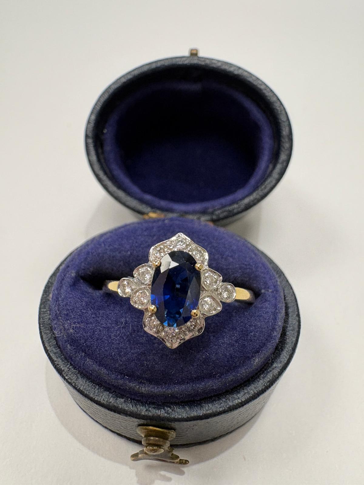 A vintage-style ring with an oval blue sapphire and diamonds in an intricate halo setting, displayed in an open round velvet ring box with a dark blue interior