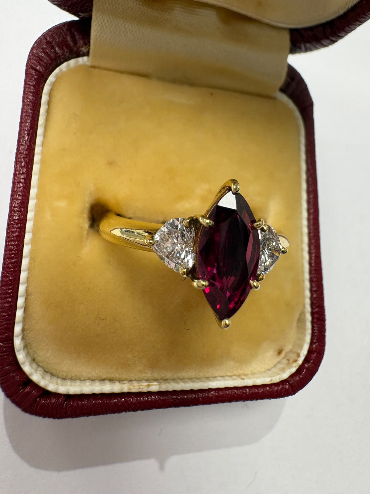 A yellow gold ring featuring a marquise-cut deep pinkish-red center stone with two small clear heart-shaped side stones, displayed inside an open burgundy and cream jewelry box.