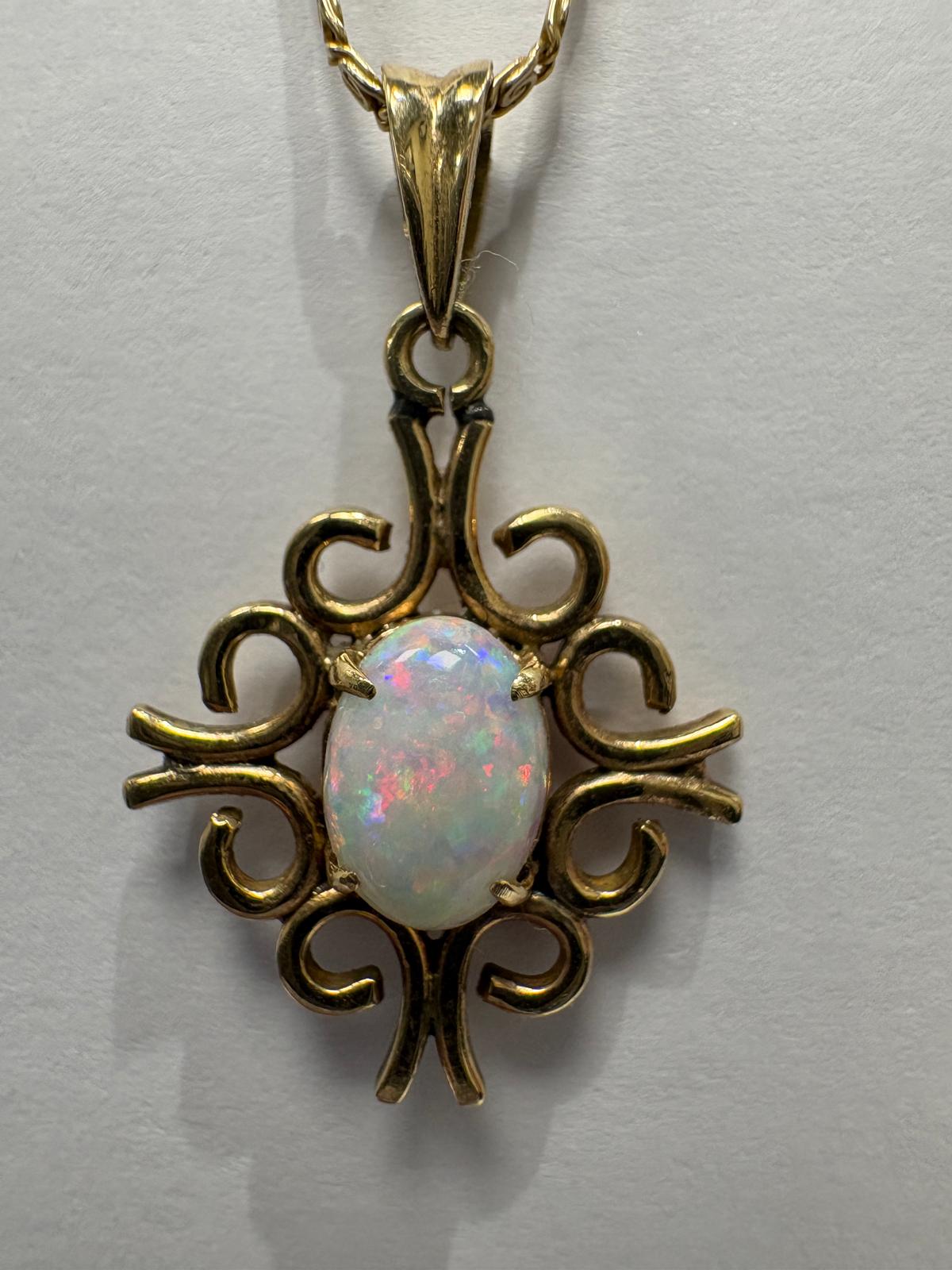 The 10k yellow gold fancy link chain leads to a 14ct yellow gold open scrollwork pendant set with an oval opal cabochon displaying white body colour with flashes of pink, red, and blue play-of-colour.
