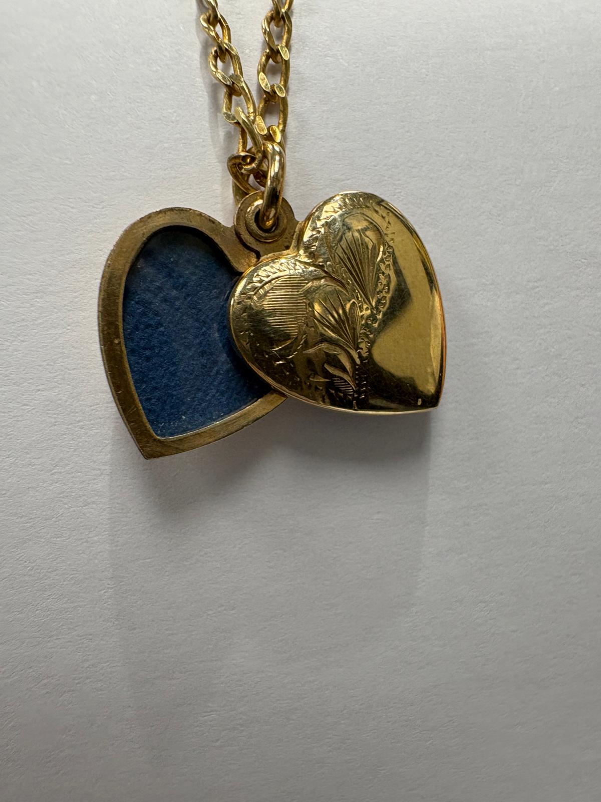 Close-up view of the vintage 9ct yellow gold heart locket open, revealing the interior. The locket opens to show a heart-shaped recess lined with blue fabric, designed to hold a photograph or keepsake. The engraved front face is visible alongside the open back panel.