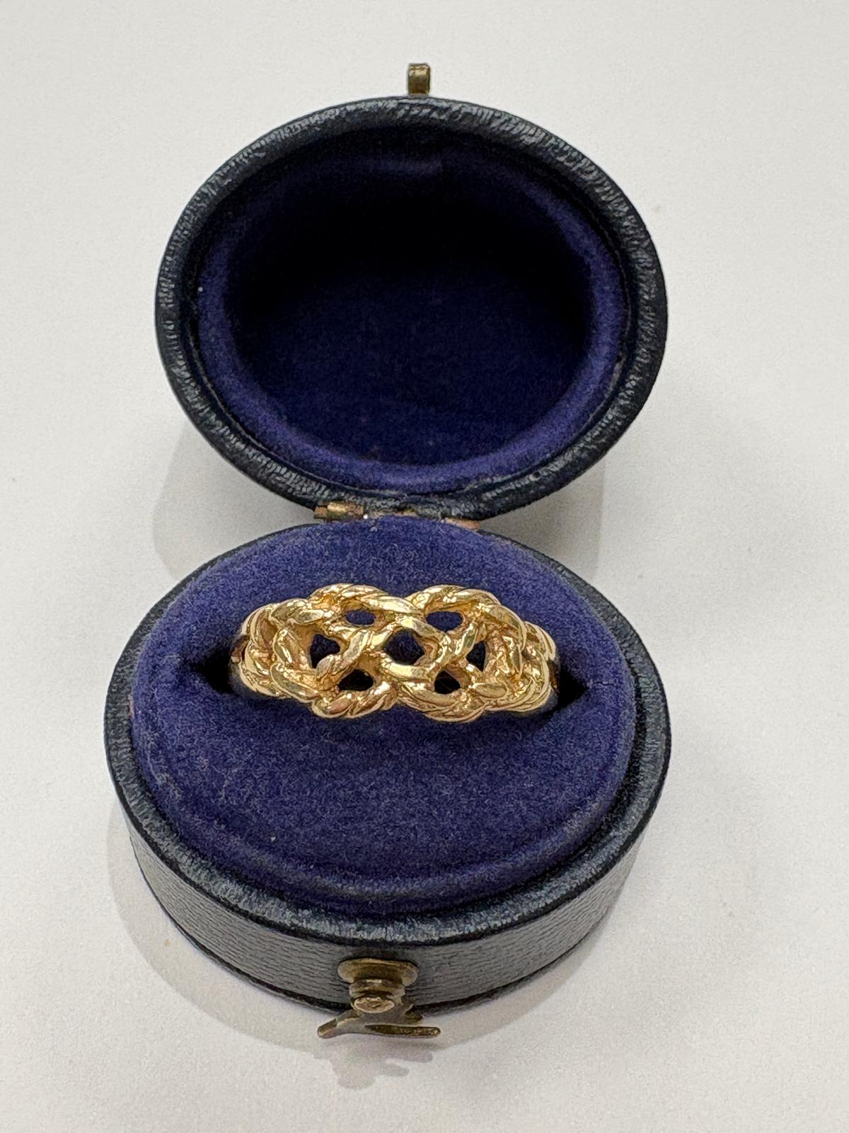 A gold ring with an intricate openwork knot pattern, displayed inside an open round black presentation box with a deep purple velvet interior.