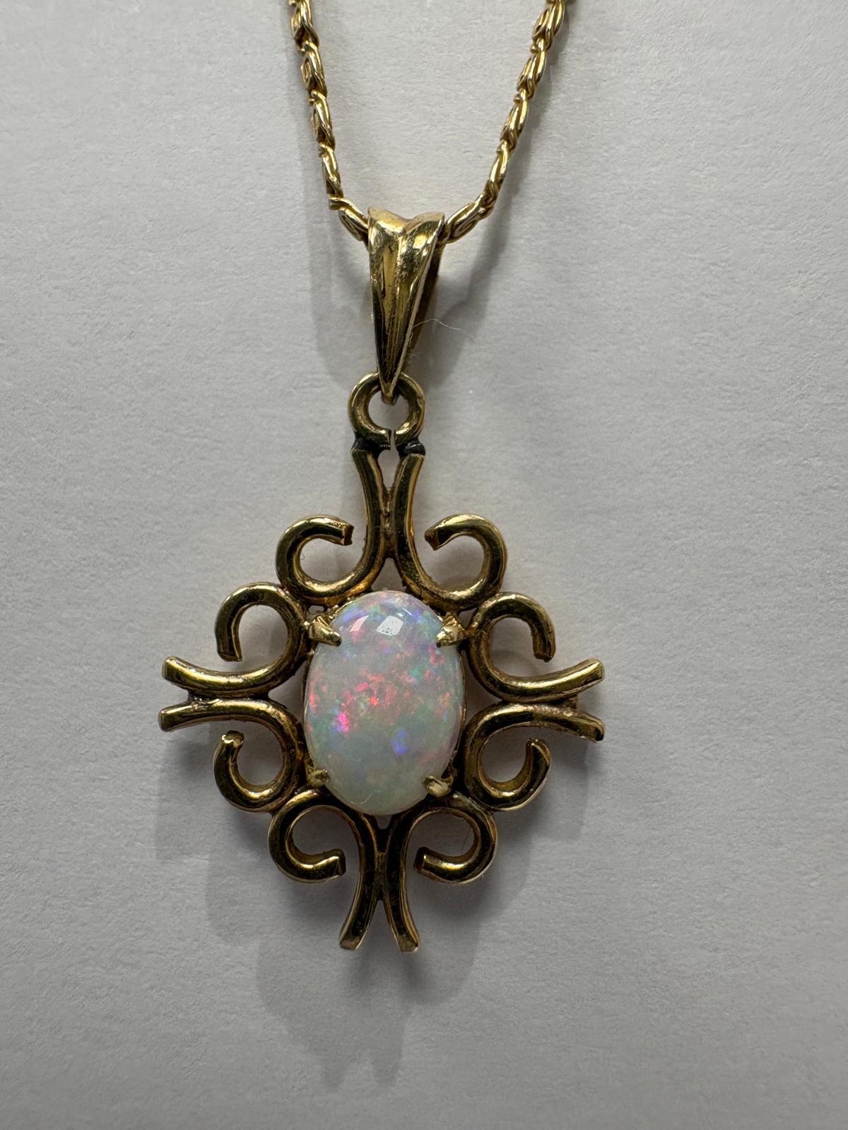 Close-up detail of a 14ct yellow gold pendant with an ornate open scrollwork frame featuring symmetrical curling motifs. The centre is set with an oval opal cabochon displaying vivid play-of-colour in pink, red, green, and blue. The opal is held by four small claw settings. A 10k yellow gold fancy link chain is visible at the top.