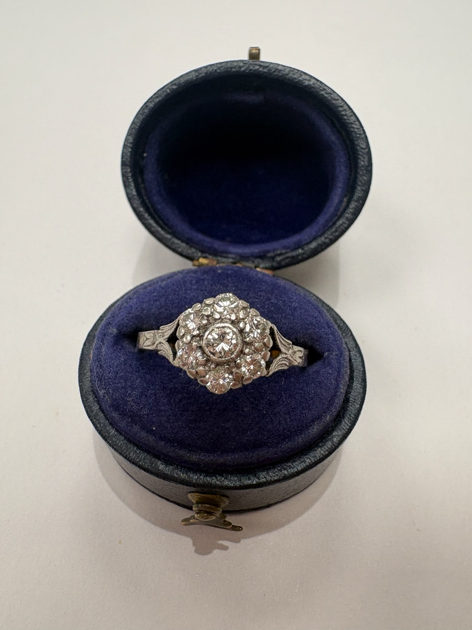 A vintage-style diamond cluster ring in a silver-toned setting, displayed inside an open round velvet jewelry box with a dark blue interior on a light background.