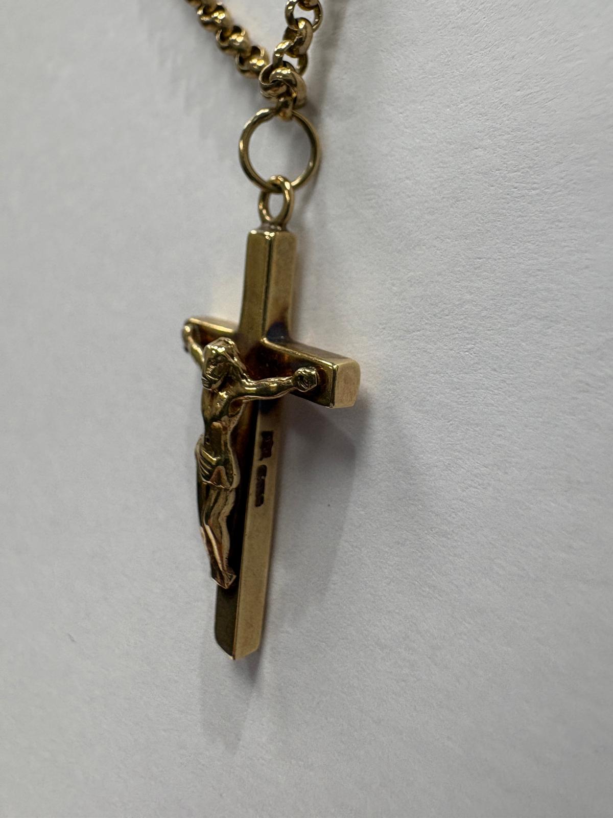 Close-up angled view of a yellow gold crucifix pendant showing the detailed corpus of Christ figure from the front left. The bevelled edges of the polished gold cross are visible, along with hallmark stamps on the lower vertical bar of the cross. The jump ring and belcher chain connection are visible at the top.