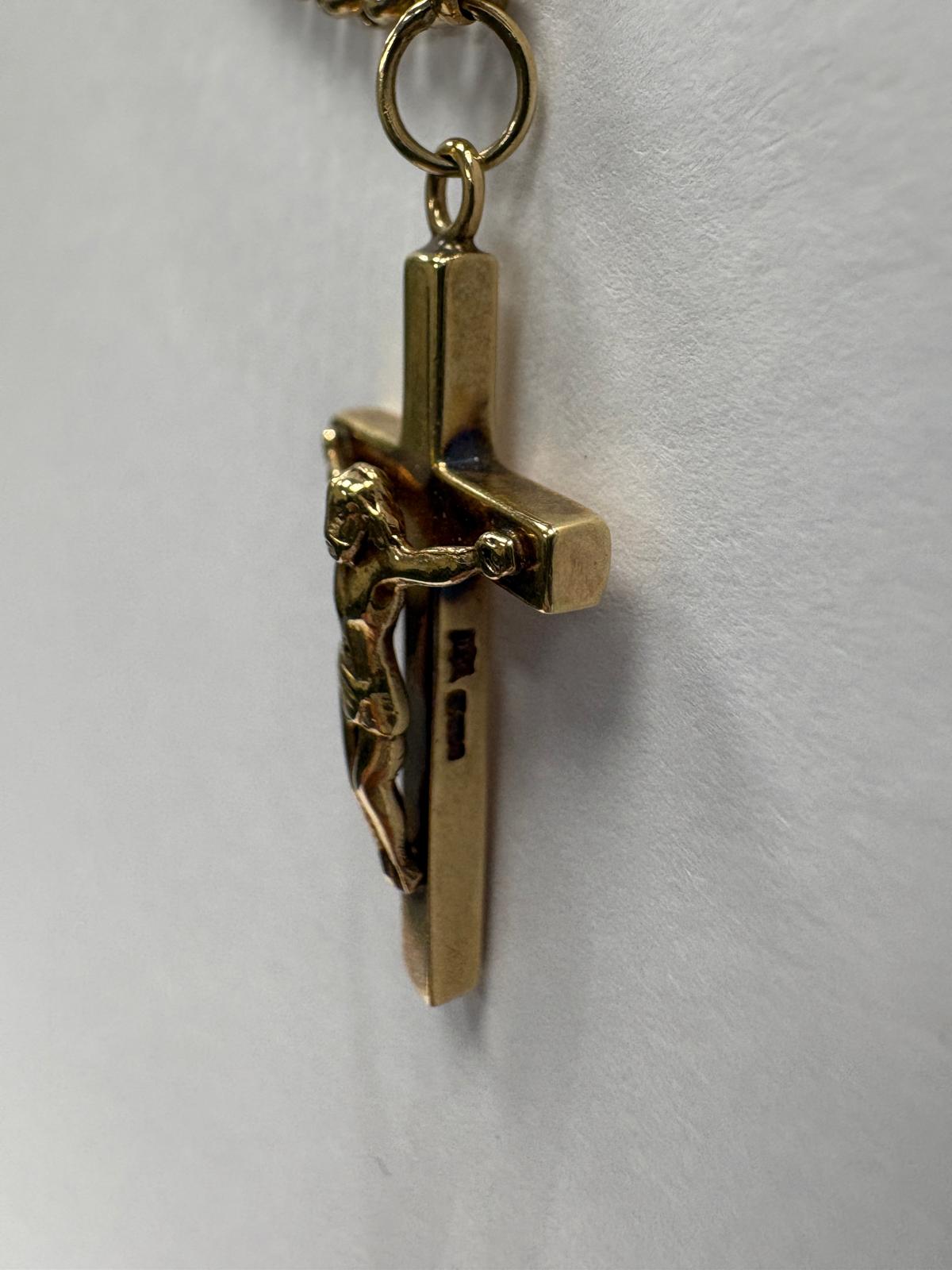 Close-up side angled view of a yellow gold crucifix pendant showing the depth and profile of the cross. The corpus of Christ is visible from the side, and hallmark stamps can be seen on the lower vertical bar. The jump ring connecting the pendant to the belcher chain is visible at the top.